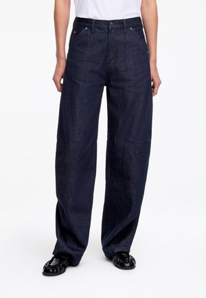 Jean en denim bleu foncé à jambes larges, taille haute, avec poches latérales et fermeture avant par boutons, porté avec des chaussures plates noires et un haut blanc.