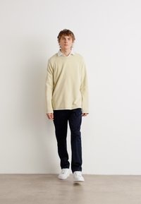 Lys beige genser med rund hals, kombinert med mørk marineblå bukser og hvite joggesko. Genseren har raglanermer og en avslappet passform.