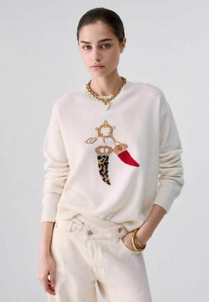 Jonge vrouw met naar achteren gekamd haar draagt een witte trui met kralenbedel design en crème broek, aangevuld met een gouden ketting en armbanden.