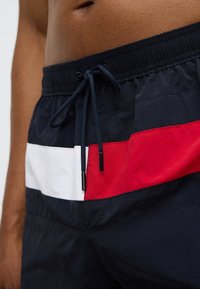 Μπλε ναυτικού χρώματος μαγιό βερμούδα με υφή, που διαθέτει κόκκινες και λευκές οριζόντιες ρίγες, ελαστική μέση και κορδόνι με μεταλλικές άκρες.