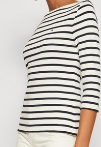 Pasiasta bluzka w kolorze kremowym i czarnym, 3/4 rękawy, okrągły dekolt, miękka tkanina, haftowany detal z logo na piersi. Klasyczny, dopasowany krój.