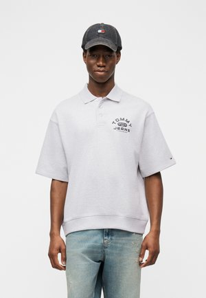 Giovane uomo che indossa una polo grigia Tommy Jeans, jeans azzurri sbiaditi e un berretto Tommy Hilfiger grigio scuro, in piedi contro uno sfondo bianco.