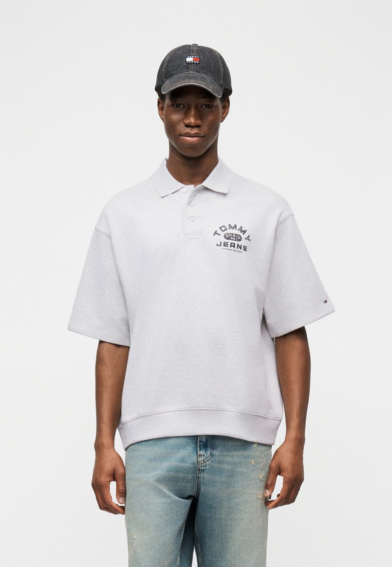 Νέος άνδρας που φοράει γκρι πόλο μπλούζα Tommy Jeans, ξεβαμμένο μπλε τζιν και σκουρόγριφο καπέλο Tommy Hilfiger, στέκεται μπροστά σε λευκό φόντο.