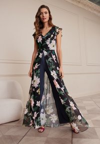 Robe maxi florale noire avec tissu transparent superposé et décolleté en V profond. Présente des manches volantées et une fente à l'avant. Associée à des sandales roses.