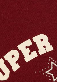 Marooni puuvillakangas, jossa on voimakasta, vaaleaa kermaista tekstiä "SUPER" koristeellisella valkoisella ompelulla ja tähden koristeilla.