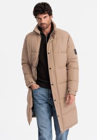 Abrigo tipo puffer en color beige con un cuello alto, botones a presión y bolsillos laterales. Fabricado con tela acolchada, presenta un detalle en parche en la manga.