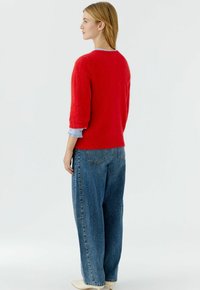 Felrood, gestructureerd truitje met driekwart mouwen, gecombineerd met blauwe denim wijde broekspijpen, gezien van achteren.