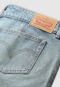 Lichtblauwe denimstof met een kenmerkend ontwerp van de achterzak en een tanleren label met rode tekst en grafische elementen.