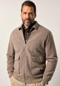Mand med skæg iført beige zip-up jakke over mørk skjorte, hænderne i lommen, står mod en lys neutral baggrund.