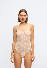 Hautfarbener Spitzen-Bodysuit mit floralem Muster, verstellbaren Trägern und gewellten Kanten. Transparenter Stoff mit hochgeschnittenem Beinausschnitt.