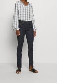 Blouse à carreaux bleu clair et blanc avec un décolleté en V et des manches bouffantes, associée à un jean skinny en denim noir avec des fentes sur le côté. Sandales noires.