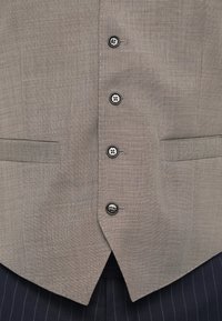 Vest griț cu un model de tip herringbone, având șase nasturi negri, două buzunare laterale și un tiv ascuțit, purtat peste pantaloni în dungi închise.