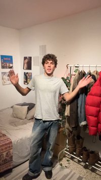 Jeune homme aux cheveux bouclés, portant un t-shirt gris et un jean, debout les bras levés dans une chambre avec des vêtements sur un portant et des affiches sur les murs.