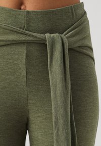 Gros plan d'une personne portant un pantalon taille haute vert olive avec une ceinture en tissu nouée à la taille.