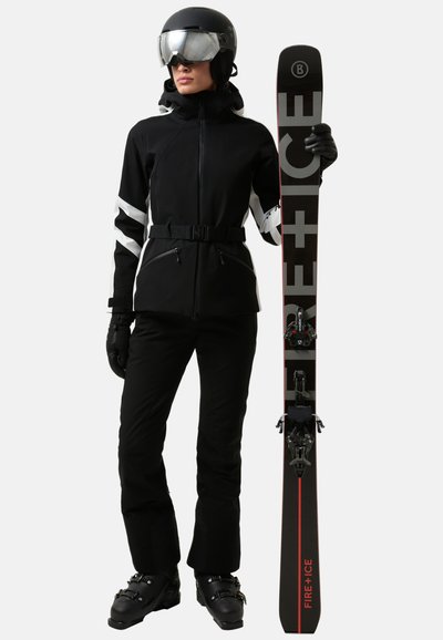 Schwarze Skijacke mit weißen Akzenten, schwarze Skihose, schwarzer Helm und Skibrille. Hält schwarze und rote Ski mit deutlich sichtbaren Bindungen.
