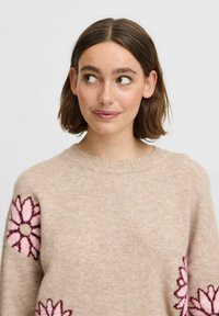 Pull en beige avec un col rond et des manches longues, orné de broderies florales roses sur les deux manches, fabriqué en tissu doux et texturé.