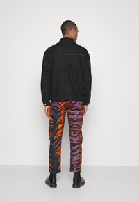 Giacca di denim nera con colletto a punta, abbinata a pantaloni con motivo astratto multicolore in arancione, viola e nero. Stivali neri alla caviglia.