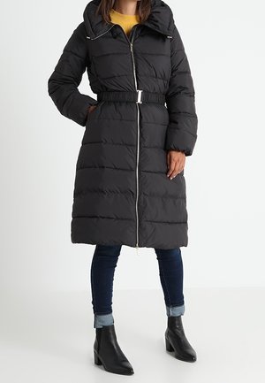 Femme portant un long manteau matelassé noir avec capuche, fermeture éclair devant, taille ceinturée, jean foncé retroussé et bottines noires à talons.
