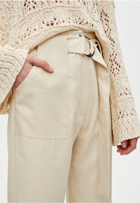 Pantalon sur mesure couleur crème avec une large ceinture dotée d'une boucle métallique argentée, poches avant, et une texture lisse. Pull beige en tricot visible.