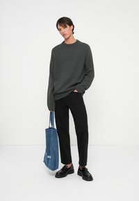 Szary dzianinowy sweter, czarne spodnie i czarne buty. Trzyma niebieską torbę denimową z napisem "SAR". Prosty, luźny krój.