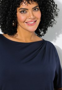 Žena s kudrnatými černými vlasy, oblečená v námořnické modré bluze a zlatými náušnicemi, se usmívá na pozadí světle šedé textury.