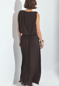 Mustapleated maxi-mekko, jossa on röyhelörintamus ja hihaton muotoilu. Kangas on pehmeää ja virtaavaa. Kultainen rannerenkauskoristus.