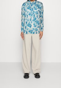 Personne portant un blouse fleuri bleu et blanc à manches longues, un pantalon large beige et des chaussures à plateforme noires, se tenant contre un fond blanc.