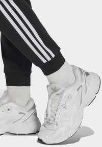 Sneakers atletici bianchi con accenti argento, upper in mesh e sintetico, punta rotonda e fondo testurizzato, abbinati a pantaloni jogger neri con strisce bianche.