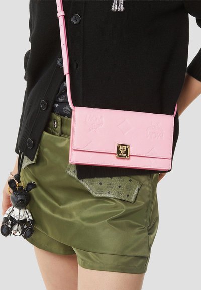 Άτομο που φοράει μαύρη ζακέτα και φούστα ελιάς με ροζ τσάντα ώμου MCM και ένα μικρό μαύρο-άσπρο μπρελόκ αρκούδας που κρέμεται από τη μέση.