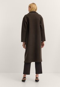 Langer, dunkelbrauner Mantel mit breitem Revers und herabfallenden Schultern, gefertigt aus strukturiertem Stoff. Verfügt über einen geraden Schnitt und minimale Verzierungen.