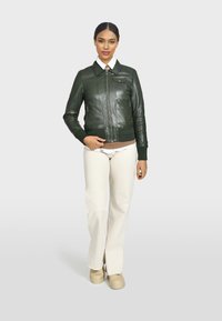 Giacca di pelle verde scuro con zip frontale, polsini e collo a coste, abbinata a pantaloni color crema e scarpe beige.