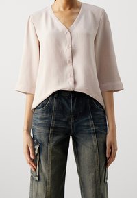 Blusa button-up rosa chiaro con scollo a V, maniche a tre quarti, tessuto testurizzato, abbinata a jeans scuri in stile cargo.