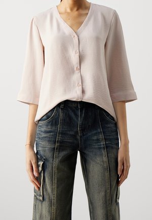 Chemisier rose clair à col en V, manches trois-quarts, en tissu texturé, associé à un jean cargo bleu foncé.