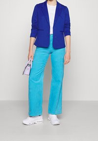Blazer azul sobre una camiseta blanca, combinado con pantalones de pana azul claros y zapatillas blancas. Pequeño bolso púrpura agarrado con la mano izquierda.