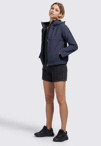 Marine waterdichte jas met een capuchon, een ritssluiting aan de voorkant en twee borstzakken, gecombineerd met zwarte shorts en zwarte sneakers. Soepele textuur.