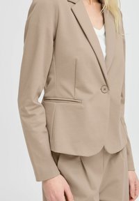 Béžový přizpůsobený blazer s jedním knoflíkem, drsně vykrojenými klopami a bočními kapsami, vyrobený z hladké, texturované tkaniny.