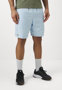 Svetlo modri šortsi iz gladkega materiala, z elastičnim pasom in zelenim logotipom Lacoste. Oblečeni z črnimi supergami in belimi nogavicami.