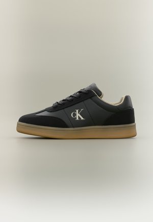 Baskets noires Calvin Klein avec semelle beige, design à lacets et logo blanc "cK" sur le côté, présentées sur fond neutre.