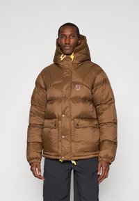 Brown puffer jacket with a hood, yellow drawstrings, two front pockets, and logo patch. Shiny fabric with a quilted texture.