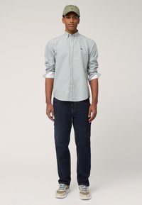 Chemise à rayures vert clair et blanche, associée à un jean bleu foncé et des baskets beiges. Complétée par une casquette vert olive.