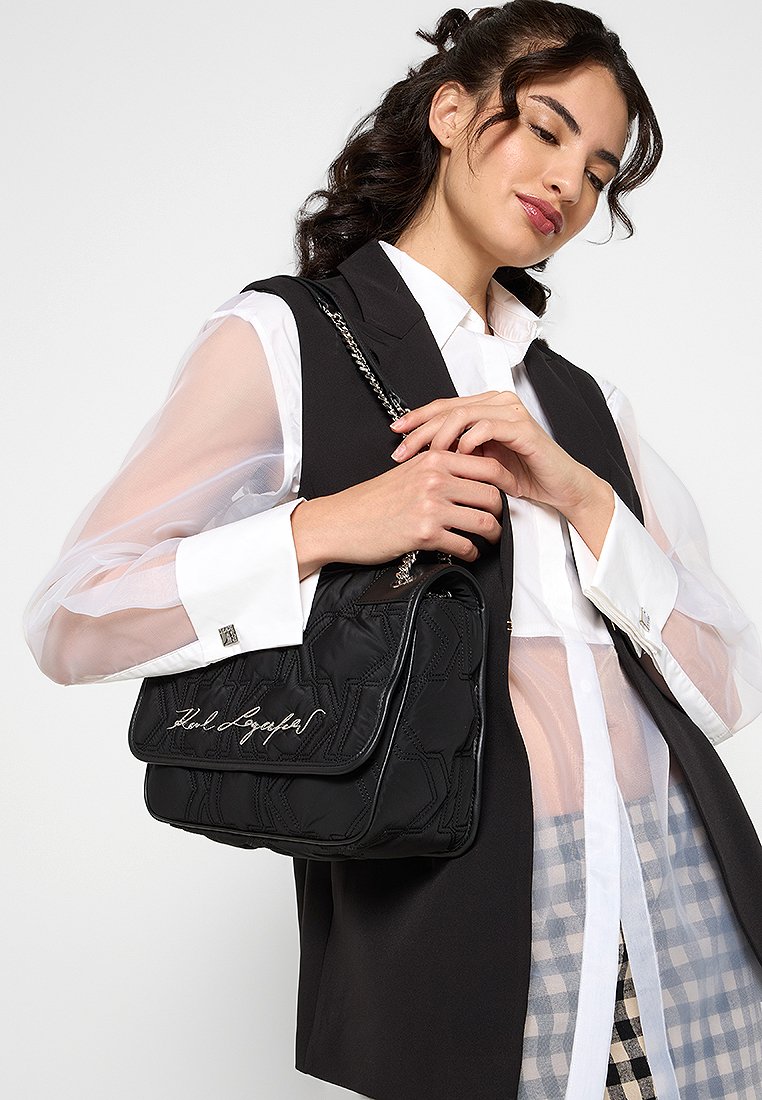 Sac à main noir matelassé avec un rabat et une bandoulière en chaîne, présentant des motifs géométriques en relief et une signature en blanc.