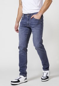 Jeans ajustados de color azul hechos de denim, con un diseño estándar de cinco bolsillos, un sutil desgaste y cierre con cremallera. Combinados con zapatillas negras y blancas.