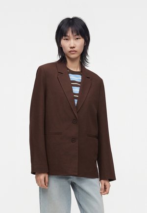 Jeune femme aux cheveux courts noirs porte un blazer marron oversize, une chemise rayée et un jean bleu clair sur un fond blanc uni.