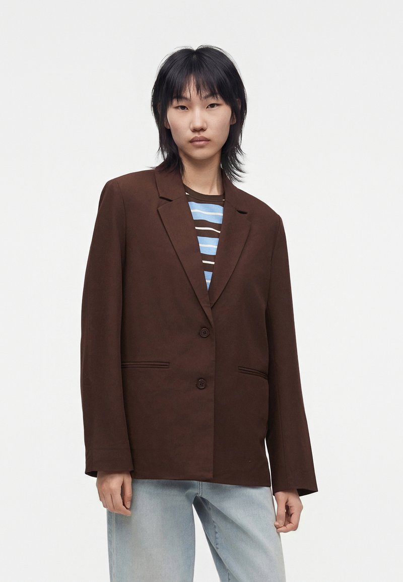 Jeune femme aux cheveux courts noirs porte un blazer marron oversize, une chemise rayée et un jean bleu clair sur un fond blanc uni.