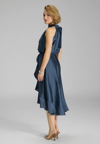 Robe en satin bleu foncé avec un col haut noué, taille cintrée et un ourlet évasé en couches. Portée avec des talons transparents. Texture lisse.