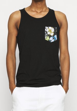 Person iført sort ærmeløs skjorte med blomsterlommedesign og hvide bukser, hænder hvilende i lommer, neutral baggrund.