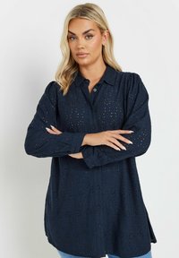Camicia blu navy con motivo ad occhielli intricato, maniche lunghe, vestibilità rilassata e orlo leggermente curvato. Dettaglio del tessuto testurizzato visibile.