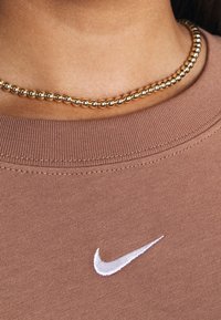 Brązowa bluza z okrągłym dekoltem, z białym logo Nike. Widać także złotą naszyjnik choker, który dodaje kontrastujący detal.