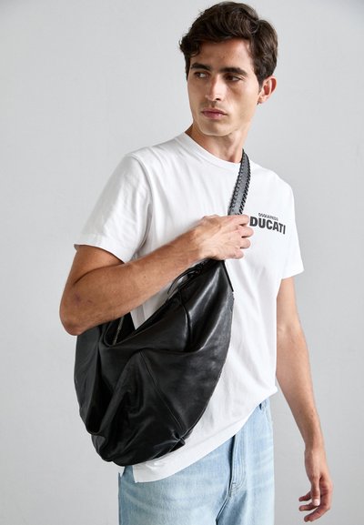Crna kožna hobo torba s mekim teksturom, zatvaranjem na zip, i zakrivljenim oblikom, drži je osoba u bijelom majici i svijetloplavim trapericama.