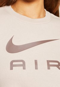Beige sweatshirt med en stor brun Nike swoosh-logotyp och texten "AIR" på framsidan. Mjuk, texturerad tyg med rundad halsringning.